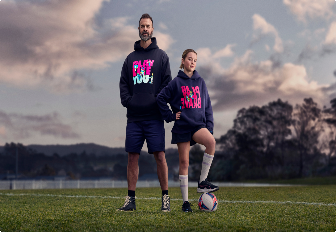 Image of a girl playing football wearing the Play Brave hoodie and her father supporting her wearing the Play Like You hoodie
