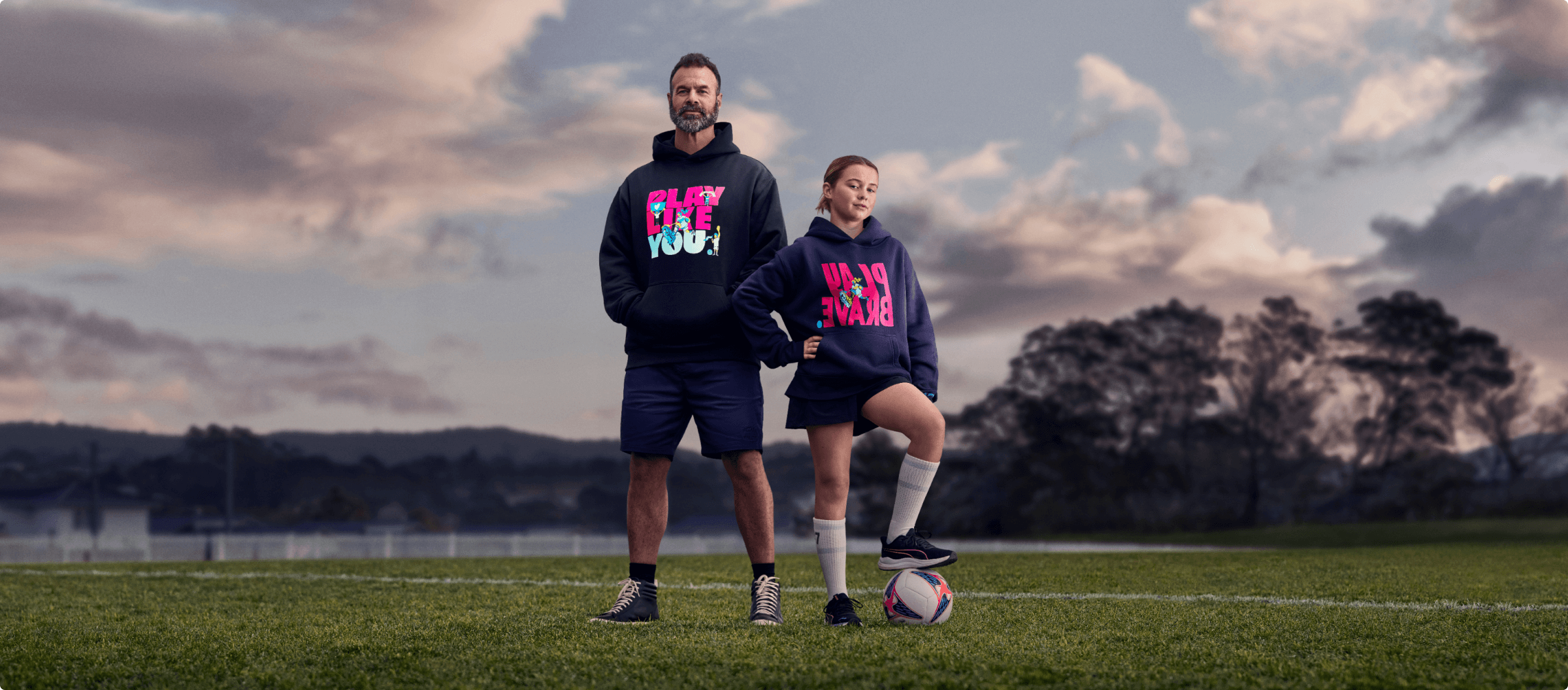 Image of a girl playing football wearing the Play Brave hoodie and her father supporting her wearing the Play Like You hoodie