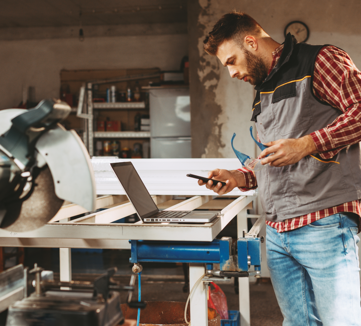 Man working in a wood workshop with a laptop, smartphone and power tools