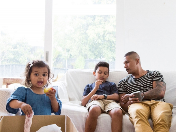 Man sitting on couch with son and daughter unpacking a box while eating an apple