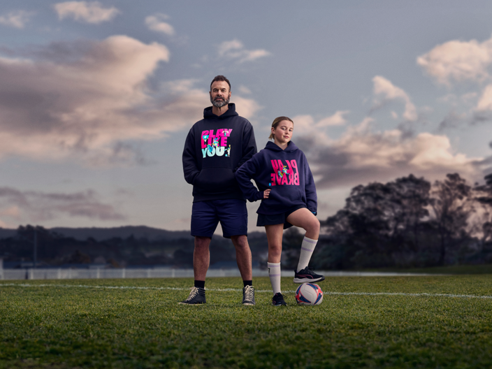 Image of a girl playing football wearing the Play Brave hoodie and her father supporting her wearing the Play Like You hoodie